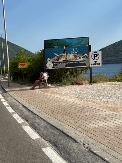 The image captures a scene on the outskirts of Perast, Montenegro, a town situated on the Bay of Kotor. In the foreground, a road with white lane markings leads towards the left of the frame. Adjacent to the road is a paved walkway made of brick-like tiles. Beyond the walkway, a person is seated on a plastic chair, leaning their head against their hand, appearing to be looking at a mobile phone. The person is wearing a white t-shirt and dark shorts. To the right of the person, a red umbrella lies on the ground.
Further into the scene, a large billboard displays an advertisement for "Hotel & Restaurant Conte, Perast" and features an image of a spread of seafood and wine, with a scenic view of mountains and water in the background. To the right of the billboard, a white sign with a blue circle and a white "P" indicates "PAY PARKING". Behind the billboard and parking sign, lush green trees and bushes are visible, and beyond that, the blue waters of the Bay of Kotor are clearly seen, with hills or mountains forming the backdrop. The sky is a clear, bright blue, suggesting a sunny day. A yellow road sign with the word "Perast" is mounted on a pole to the left of the billboard.