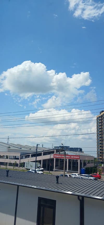 The image depicts a view of Novaya Adygeya, Russia, on a sunny day with a bright blue sky filled with cumulus clouds. In the foreground, a white building with a dark gray corrugated metal roof is visible, featuring two black vent pipes. Beyond this building, a multi-story parking garage with a pink facade is situated. A red banner on the parking garage reads "ПРОДАЖА ПАРКОВОК 8 861 205-45-57", which translates to "PARKING SALES". Several cars are parked in front of the parking garage. Further in the background, a tall residential building with numerous windows and balconies stands to the right, and other apartment buildings are visible behind the parking garage. Utility poles and overhead power lines traverse the middle ground of the image. The overall scene suggests an urban environment on a clear day.
