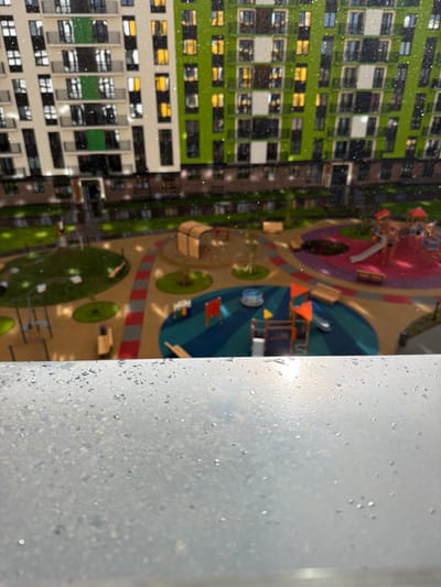 The image depicts a view from a window with water droplets on the glass, looking out onto a modern residential courtyard in Pervomaysky, Russia. In the background, a multi-story apartment building with a facade of white, green, and beige panels and numerous windows is visible. The courtyard features a playground with slides, climbing structures, and swings, all set on a colorful, patterned rubberized surface. There are also grassy areas, benches, and a sandpit with a wooden arched structure. The ground is wet, suggesting recent rain. The overall scene is one of urban residential infrastructure in what appears to be inclement weather.