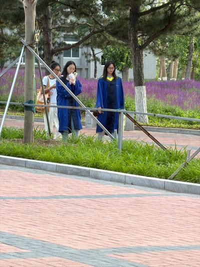 Three young women, appearing to be students, stand outdoors in what seems to be a park or campus setting. The woman on the left wears glasses and a white t-shirt with light-colored pants, carrying a tan tote bag with a camera attached. The woman in the middle wears glasses and a blue academic gown over a white shirt and jeans, holding a pink beverage and her phone. The woman on the right also wears a blue academic gown over a yellow collared shirt and jeans. They are positioned in front of a blurred background of buildings and a field of purple flowers. The foreground features a paved walkway with red and grey brickwork. The scene is likely in Guta, China, given the context of the provided location.
