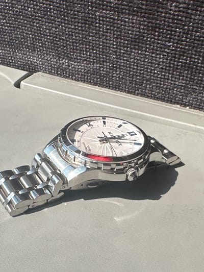A metallic wristwatch is observed resting on a flat, light gray surface under direct, strong illumination from the right, casting a prominent shadow. The watch features a silver-colored case, a three-link metal bracelet, and a textured, silver-toned bezel. Its light-colored dial displays a radial pattern, along with Roman numerals (e.g., XII) and a 24-hour track marked with Arabic numerals. The text "ICE OUT" and "EXCLUSIVE TOP" is visible on the dial. A red band encircles the dial circumference beneath the bezel. A knurled crown is present on the right side of the watch case. The background consists of a dark, out-of-focus, textured material, separated from the foreground by a raised, light gray horizontal edge. No individuals or active subjects are present. The scene is located in Gorna Vasilitsa, Bulgaria.