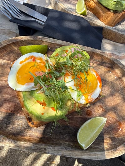This is a close-up, overhead shot of an avocado toast with two fried eggs served on a rustic wooden board. The toast is topped with sliced avocado, two perfectly cooked fried eggs with runny yolks, and a garnish of fresh microgreens. A slice of lime is placed to the side of the toast, and another lime wedge is visible to the left of the frame. A dark napkin and a fork and knife are also present on the board, suggesting the meal is ready to be eaten. The background shows a sandy surface and the dappled sunlight indicates it is daytime, likely outdoors. The overall impression is a healthy and appealing breakfast or brunch. The setting appears to be a casual outdoor dining area, potentially a beach cafe, given the sand. The sunlight casting shadows on the wooden board suggests a sunny day. There is no visible text in the image.