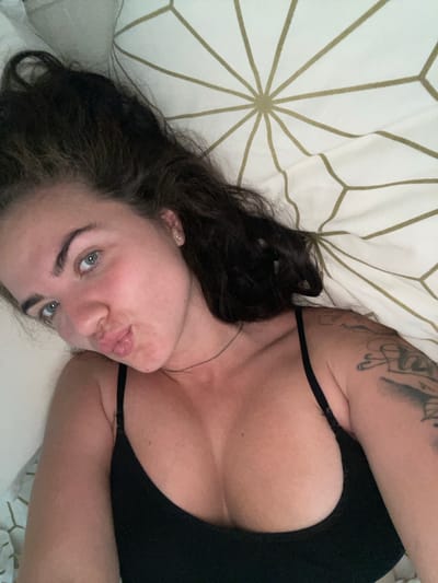 A close-up, overhead shot shows a woman lying in bed. Her face is angled towards the camera, with her lips pursed. She has light blue eyes and dark brown hair. She is wearing a black camisole, and a portion of a tattoo is visible on her right arm. The background is a white bedsheet with a repeating geometric pattern in a light gold color. There is no information in the image to determine the location within the city of Мосметаллоконструкция Онт, Russia.