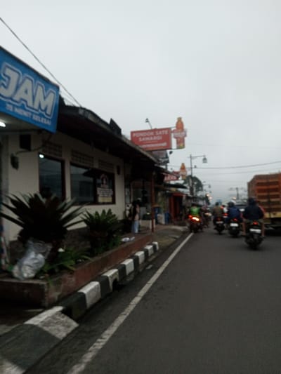 This image captures an urban street scene in Chagzam_Pam, Bhutan, under an overcast sky. On the left, a light-colored building features a partial blue sign reading "JAM" and "TO MENIT SELESA." Further along the sidewalk, a red sign advertises "PONDOK SATE SAWARGI" with an attached beverage bottle advertisement. Dark green plants are visible in a planter box along the sidewalk, near which a white plastic bag rests. A single individual stands on the sidewalk adjacent to the building. A black and white striped curb separates the sidewalk from the asphalt road, marked with a solid white line. On the road, several motorcyclists, most wearing helmets, are traveling away from the viewer. A large brown truck is also visible in the traffic. Overhead, power lines, utility poles, and street lights are discernible against the gray sky.