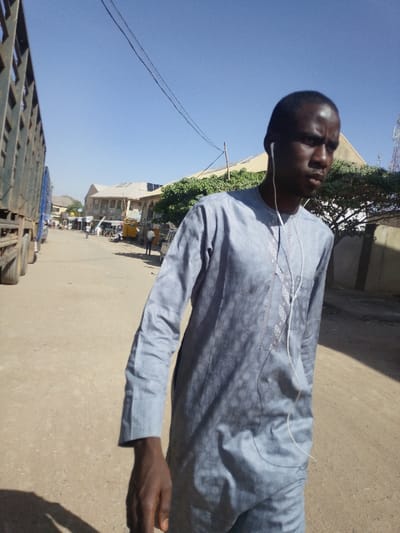 An adult male is depicted walking outdoors on an unpaved road in Babban Giji, Nigeria, under a clear blue sky. The subject, of dark complexion, wears a light grey or blue long-sleeved tunic with subtle patterned designs and wired white earbuds. He looks towards the right, out of the frame. To the left, several large, dark-colored trucks with slatted sides are parked or slowly moving along the dusty road. In the background, a street scene unfolds with multiple single and multi-story buildings, some with corrugated metal roofs, suggesting commercial or residential use. Green foliage from trees is visible on the right, and overhead power lines traverse the sky. Distant figures are visible further down the road, and a red cellular tower is present on the far right horizon. The scene is brightly lit by natural sunlight, casting visible shadows.