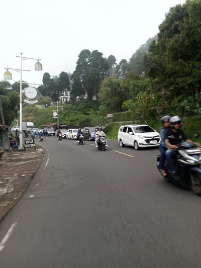The image depicts a street scene in a mountainous region. The road is lined with trees and vegetation. There are several vehicles on the road, including motorcycles and cars. People are riding motorcycles. A sign that says "VILA KAMAR" is seen on the left side of the frame. There are light posts on the left side of the road, adorned with Arabic writing. The sky is overcast. The location appears to be somewhere in Southeast Asia.