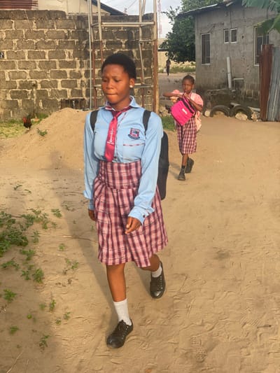 The image depicts two female students walking on a dirt path in Mosafejaw, Nigeria. The foreground features an adolescent female, with dark skin and short dark hair, wearing a light blue long-sleeved school shirt with a crest on the left pocket, a red tie, a red and white plaid skirt, light grey socks, and black lace-up shoes. She carries a black backpack and walks with her head slightly down, looking forward. To her right and further back, a younger female child, also with dark skin and dark hair, wears a light pink short-sleeved top, a matching plaid skirt, and black shoes. She carries a bright pink bag in her left hand and looks towards the viewer.
The environment includes a rough concrete block wall on the left, with a pile of sand or dirt and sparse green vegetation. A dark-feathered fowl stands on the dirt pile. Behind this wall, a metal structure is visible. On the right, a grey building with small windows stands, with discarded tires and corrugated metal sheets near its base. In the far background, a faint figure walks on a distant path, and a telecommunications mast is visible against the sky. Green foliage and trees are also present in the background. The ground is a light-colored sandy dirt path with visible footprints. The scene is illuminated by natural daylight.