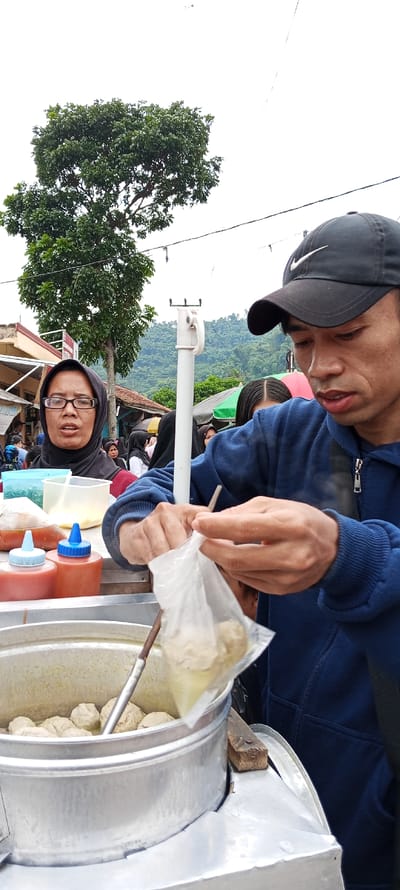 A man in a blue hoodie and a black baseball cap is picking up a serving of food from a steaming pot. The food is round and white, and it is being served in a plastic bag. The man is standing next to a food vendor, who is wearing a headscarf. The vendor is standing behind a cart filled with supplies, including a metal pot, a wooden board, and bottles of red sauce. The cart is located in the city of Kecamatan Kutawaringin, Indonesia, near a street with a large tree and hills in the background. The scene is sunny and bright. The man appears to be purchasing food from the vendor.