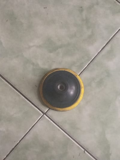 The image displays a circular, disk-shaped object resting on a tiled floor. The object features a bright yellow outer rim and a dark gray or black central section. In the center of the dark area, there is a circular indentation with a visible threaded hole, indicating it is likely an attachment point for a power tool, possibly a sanding, buffing, or backing pad. The object appears to be used, showing some minor wear or discoloration, particularly on the yellow rim.
The setting is an indoor space, with the object placed on a tiled floor. The tiles are a light, muted green or gray-green color, arranged in a grid pattern with darker grout lines. There are no people present, and no activity is depicted. The lighting is neutral, suggesting an indoor environment. No text is visible on the object or in the surrounding environment. The scene is located in Kecamatan Arjasa, Indonesia.