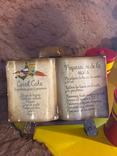 This image depicts a wooden carving resembling an open recipe book, situated indoors. The book's pages feature recipes in Spanish. The left page has an illustration of a Carrot Cake, along with a list of ingredients intended for six people, including flour, grated carrots, eggs, oil, peeled nuts, and cinnamon. The right page contains instructions titled "Preparación de la receta" (Preparation of the recipe), detailing how to mix the ingredients, bake at 150°C for 60 minutes, and decorate with nuts. The carving appears to be part of a larger decorative structure, as suggested by the visible portion of a yellow and red object in the background. The exact location in San Martín de la Vega, Spain, is impossible to determine from the image alone.