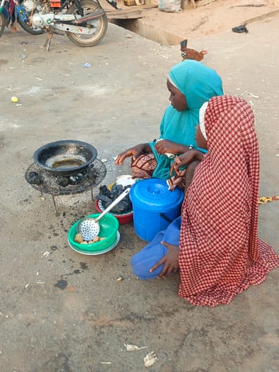 The image portrays an outdoor scene in Alizaga, Nigeria, featuring two individuals seated on the ground alongside a cooking setup. A black, round cooking pot containing a liquid, likely oil, rests on a metal charcoal grill with visible embers. Adjacent to the grill, a green plastic colander holds multiple light-brown, cuboid food items, with a white metal slotted spoon placed inside it. A blue lidded plastic bucket and a red bowl containing dark objects are situated next to the colander.
One individual, wearing a turquoise head covering and garment, is seated on the left, facing towards the cooking area with hands near the food items. The second individual, wearing a red and white checkered head covering and garment, is seated on the right, partially facing away from the cooking setup.
In the background, the rear wheel and part of the frame of a parked motorcycle are visible on the left. A brown chicken is present in the mid-ground behind the seated individuals. The ground surface consists of uneven concrete and dirt, exhibiting scattered debris and dark stains. A concrete curb is discernible further in the background.