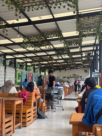 This image depicts a casual, semi-open-air gathering place, likely a restaurant or food court, in Bekasi Regency, Indonesia.

Numerous people are present, mostly seated at low wooden tables and benches constructed from pallets, arranged in rows. Some individuals are seen conversing, eating, or using their phones. In the foreground, a person wearing an orange top and a black head covering is seated on a pallet bench, with a red fabric item beside them. Another person in a blue shirt is partially visible on the right. A man in a black t-shirt and cap stands near a grey wheeled trolley, looking down. Farther down the rows, more patrons are visible, some wearing head coverings, engaged in various activities.

Overhead, a metal framework supports a roof made of corrugated panels, allowing natural light to filter through. Artificial ivy vines and small white string lights are draped across these metal beams, adding a decorative touch. Several black ceiling fans are also mounted to the structure.

Notable details include the distinctive wooden pallet furniture, which serves as both tables and seating, with the brand name "GACOAN" visible on some of the crates. In the background, a large sign reads "THE LAND OF GACOAN", suggesting this is a branch of a food establishment. The bright ambient light indicates it is daytime, possibly morning or afternoon. The overall atmosphere appears relaxed and communal.