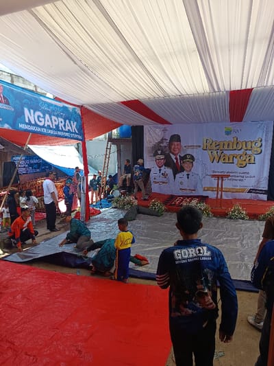 The image depicts an outdoor event taking place under a large tent structure in Kawungluwuk, Indonesia. The tent ceiling is draped with alternating white and red fabric panels. The ground is covered with a red tarpaulin in the foreground, transitioning to a grey tarpaulin further back towards a raised stage area.
On the stage, a large white banner serves as a backdrop, featuring images of three Indonesian officials, one centrally placed and two smaller ones below him. The banner displays the text "Rembug Warga" (Community Discussion) in gold, followed by a motto in Indonesian: "Bersatu dalam suara, majukan bersama untuk Cianjur istimewa" (United in voice, together advance for a special Cianjur). The stage is adorned with white floral arrangements on either side of a dark podium.
To the left of the stage, a blue banner hangs, displaying "Selamat Datang NGAPRAK" (Welcome to NGAPRAK) and "MENDAKAN KELUARGA BERISIKO STUNTING" (identifying families at risk of stunting), suggesting the event's focus on community health and development.
In the foreground and midground, approximately a dozen individuals are present. A man in a blue long-sleeved shirt with "GOROL GOYONG ROYONG LOBAAN" and a graphic on his back is seen from behind, facing the stage. Nearby, a young boy in a yellow shirt and blue pants stands. Several adults, including one in an orange long-sleeved shirt and multiple individuals in green or patterned attire, are actively kneeling and arranging or securing the grey tarpaulin on the ground. Other people are standing and observing various activities around the event space. In the background, a wooden ladder leans against a building, and a blue barrel is visible. The scene is brightly lit by natural sunlight.