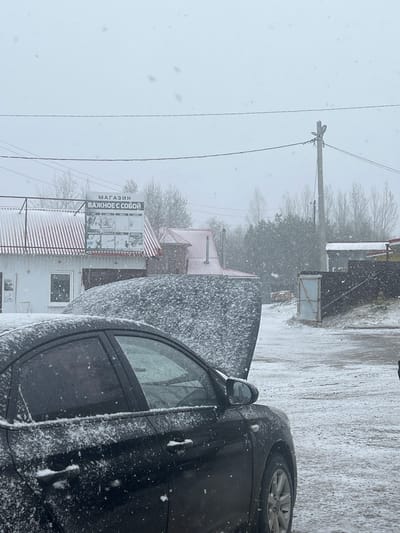 The image shows a snowy street scene in Kliuchi, Russia. A black car is parked on the right side of the frame, covered in snow. In the background, there is a building with a sign that reads "МАГАЗИН ВАЖНОЕ С СОБОЙ" (translated as "Shop Important With You"). Power lines run along the left side of the image. Snow is falling heavily, obscuring the details of the landscape.