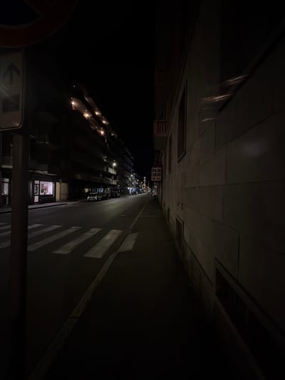 This is a nighttime, low-angle shot of a city street in Novara, Italy. The perspective is skewed, making the street appear to stretch upwards towards the viewer. The scene is dark, with artificial lights illuminating parts of the road and buildings.
On the left side of the street, there are buildings with rows of windows that emit a warm, yellow light. Some of these windows appear to be part of apartments or shops. Further down the street, a sign is visible, reading "SPAZIO LIBERO" in white text on a black background, with smaller text below.
On the right side of the street, a road is visible with pedestrian crossings marked by white stripes. Parked cars line the right side of the road, their forms mostly obscured by the darkness. Shadows are cast by the overhead structures and lights, creating a dramatic effect.
In the foreground, on the bottom left, a red and orange circular sign is partially visible. Below it, a white rectangular sign with a black arrow pointing left indicates a direction.
The overall mood of the image is one of urban quietude at night. The time of day is clearly night due to the artificial lighting and darkness. There are no people actively engaged in activities visible in the frame, contributing to a sense of stillness. The location is identified as Novara, Italy, which might suggest the architectural style of the buildings. The weather appears to be clear, as there are no visible indications of rain or fog.