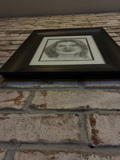 A close-up, angled photograph captures a framed pencil drawing displayed on a textured wall. The central subject is a grayscale portrait, likely of a young woman or girl, depicted with a gentle smile and her head tilted slightly. Her hair appears to be swept back or to one side. The drawing is encased in a dark-colored, possibly black or very dark brown, frame with a white mat board.
The wall exhibits distinct textures. To the left of the frame, there's a section covered with narrow, vertically oriented, light-colored bricks or tiles. To the right of the frame, the wall is composed of larger, irregularly shaped bricks or stones that are primarily light-colored, possibly painted white or light grey. These larger bricks show significant texture and wear, with hints of a darker, reddish-brown material beneath the surface, separated by visible, pronounced dark grey mortar lines.
The lighting is somewhat subdued, creating soft shadows and highlighting the various surface textures. In the upper right corner of the pencil drawing, faint, handwritten text is discernible, including what appears to be a signature or initials and the year "2019". The image portrays a static, indoor scene focused on the artwork and its immediate surroundings.