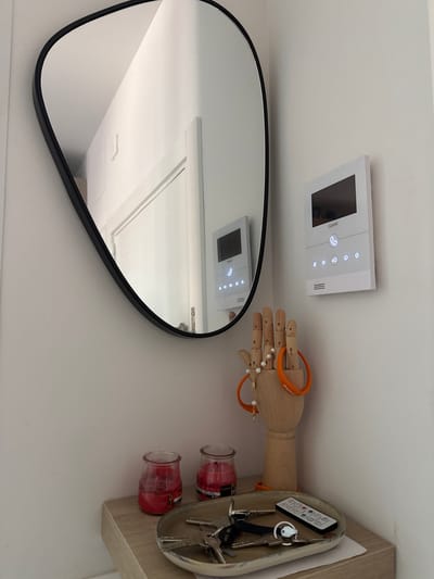 This is a medium shot of a hallway or entryway, likely in an apartment building in Getafe, Spain. The scene is viewed from a slightly elevated angle.
On the right wall, a white video intercom system with a screen and buttons is mounted. The brand "Galak" is visible on the device. To the left of the intercom, a large, irregularly shaped mirror with a black frame hangs on the wall. The mirror reflects a portion of the hallway, including a white door or wall and another, smaller intercom system, also with illuminated buttons.
On a small wooden shelf below the mirror and to the left of the intercom, two red jar candles are placed. In the center of the shelf, a wooden mannequin hand stands upright, adorned with a pearl necklace with a cross pendant and an orange bracelet that says "enjoy!". A small, silver, oval-shaped tray sits in front of the mannequin hand, containing several keys and a small white remote control with numerous buttons. The shelf itself appears to be made of light-colored wood. The walls are painted a light, neutral color, possibly white or off-white, and the ceiling is visible in the upper left of the frame.