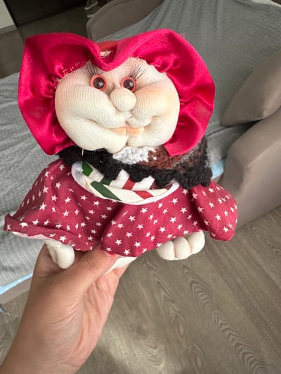A handmade doll with a red bonnet and a red dress with white stars is held by a person's hand. The doll has round plastic eyes, a bulbous nose, and a smirking mouth. It is wearing a black and white ruffled collar. The background is blurred, showing a grey sofa and a wooden floor. The image is taken indoors, in what appears to be a residential setting. No specific location in Lytkarino, Russia, can be determined from the image.
