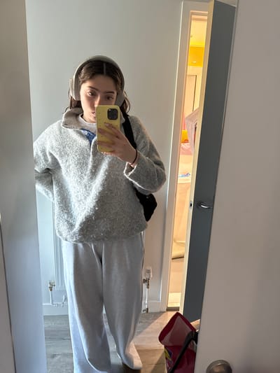A young woman is captured in a mirror selfie, holding a yellow-cased smartphone in her right hand to take the photo. She has dark hair, wears light grey over-ear headphones, and is dressed in a light grey textured cardigan or jacket over a white t-shirt, paired with matching light grey sweatpants. A black strap from what appears to be a fanny pack or crossbody bag is visible across her chest.

The setting is an indoor space, likely a hallway or bedroom. The floor is covered with light-toned wooden planks. In the bottom right corner of the mirror's reflection, a white radiator with brass piping and a valve is partially visible. Directly above the mirror, there's an open, lit storage compartment or shelf. This area emits a warm, yellow glow and contains various items, including what looks like white fabrics, yellow objects, and a red bag with the partial text "Ofo" visible. A small metal hook is also attached to the underside of the shelf's frame. The walls are plain white. The overall impression is a casual, domestic scene, typical of a home in the United Kingdom. The bright lighting suggests it is daytime.