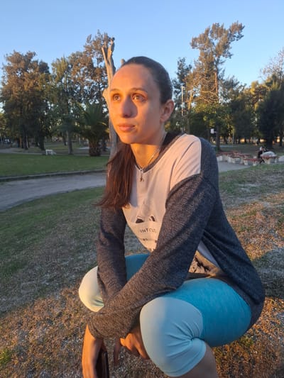 A woman in sportswear is photographed in a park in Villa Domínico, Argentina. She wears a long-sleeved grey and white shirt and blue shorts. She is crouched or bending over, looking towards the camera. In the background, there is a paved pathway with benches and trees. The environment appears to be a public park or cemetery. The sky is visible in the background.