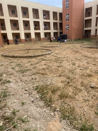 The image captures an outdoor courtyard scene in Baganakwo, Nigeria, fronting a multi-story educational building identified by the text "CENTRAL CLASSROOMS" on its facade. The ground consists of dry, light brown soil interspersed with sparse, dried vegetation, green weeds, and scattered debris including small stones and litter. Three circular concrete planters, containing parched earth and small, leafless saplings, are visible in the foreground and mid-ground. The building, constructed with beige walls and terracotta-brown accents, features multiple floors with balconies, each equipped with brown railings and square windows. A distinct reddish-brown tower-like section with several white-framed windows is integrated into the building on the right side. Approximately 20-30 individuals are present. A group of ten people, some in traditional attire, are clustered near two large, black utility tanks located at the base of the reddish-brown building section; one individual within this group is kneeling on the ground. Additional individuals are observed walking along the ground level near the building's base, and several others are standing on the upper-floor balconies. The sky above is overcast or hazy light grey.