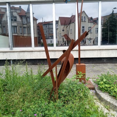 This image captures a unique outdoor scene in Eisenach, Germany, featuring a rusted metal sculpture positioned in front of a building with large, reflective windows. The sculpture is abstract, composed of several metal beams and a curved, bowl-like element, all showing signs of rust. It is surrounded by lush green vegetation, including tall grasses and flowering plants with small purple and yellow blooms.

The building's windows reflect the surrounding environment, showcasing buildings with red tile roofs and traditional European architecture. The reflections also show a cloudy sky, suggesting it's likely an overcast day. The building itself appears modern, with a band of granite-like material running beneath the windows, and a darker lower section. The ground in front of the building and sculpture is paved with square concrete tiles.

The overall impression is one of a juxtaposition between the weathered, artistic sculpture and the clean lines of the modern building, with nature actively reclaiming the space around the sculpture. There are no people clearly visible in the image, though a faint reflection of what might be a person holding a phone can be seen in the building's windows. The scene evokes a sense of quiet contemplation, with the sculpture serving as a focal point amidst the greenery and architectural reflections.