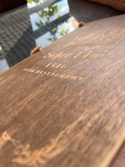 This media file shows a close-up, angled view of a wooden surface with carved text. The wood has a dark, weathered appearance with visible grain lines. The text appears to be etched or carved into the wood and is partially obscured by the angle and lighting. Some visible text includes what looks like "ESTD 1510" and "AUSTRALIA". The shallow depth of field blurs the background, which shows reflections of green foliage and what appears to be a checkered pattern, possibly a table. The lighting suggests it is daytime, with sunlight casting shadows and highlights on the wood. The overall impression is of an aged object, possibly a sign or plaque, displayed outdoors. The background hints at an outdoor setting, possibly a patio or garden area. There are also small, fibrous attachments visible on the edge of the wooden surface, possibly part of a rope or twine used for hanging.