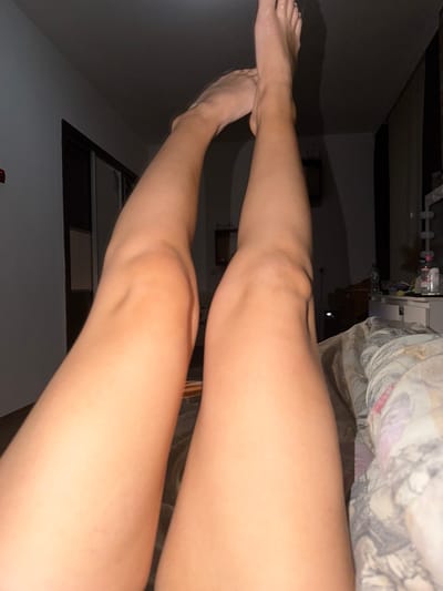 The image displays a close-up view of a person's legs, which are extended upwards. The legs appear to be tanned and are positioned against a background that suggests an indoor environment. Details of the room include a dark ceiling, a closet with sliding doors, and furniture that includes a dresser or vanity with mirrors and bottles. A patterned duvet or blanket is visible in the lower right portion of the frame. The lighting suggests it is nighttime or a dimly lit room. The image does not contain any specific identifiable landmarks or features that would indicate a location in Zmeitsa, Bulgaria.