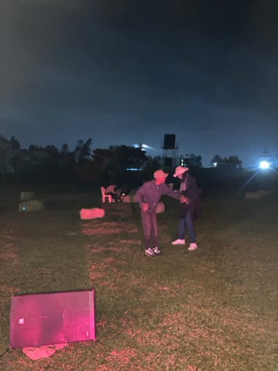 The scene depicts an outdoor environment at night in Murera ward, Kenya. A grassy field is illuminated by artificial light. In the foreground, a dark rectangular speaker rests on the grass, illuminated by a reddish-pink light. Two individuals stand in the mid-ground. The person on the left wears a light-colored cap, dark long-sleeved clothing, dark trousers, and dark athletic shoes with reflective accents. They are leaning forward with their right arm extended towards the second person. The person on the right wears a light-colored cap, a dark long coat, blue jeans, and white sneakers, facing the first individual. Their left arm is in contact with the extended arm of the first individual. Scattered across the grass in the background are two irregular dark objects and two rectangular objects resembling hay bales, all receiving reddish illumination. Further back, two red plastic chairs and a table are visible, with one person partially seated. The background features a line of dark trees, above which a metal structure with a dark water tank is visible on the right. Distant bright white lights emanate from the right side of the frame and from a large structure in the central background. The sky is dark.