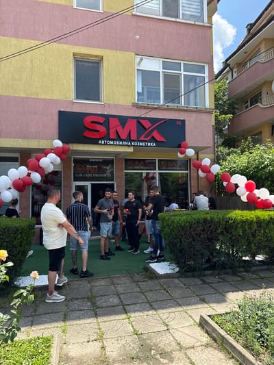 This image captures a daytime scene outside a commercial establishment located in Targovishte, Bulgaria. The central focus is a two-story building with a multi-toned facade, featuring horizontal bands of textured pink and yellow plaster. The ground floor houses a store named "SMX Автомобилна Козметика" (SMX Automotive Cosmetics), prominently displayed on a black rectangular sign with red raised lettering. Below the Cyrillic text, the English translation "CAR CARE PRODUCTS" is visible on window signage.
The storefront is decorated with numerous red and white balloons, forming garlands along the top edge of the windows and grouped in clusters to the left and right, indicating a festive event such as an opening or promotion. The entrance is open, revealing more people inside and a display of products.
A gathering of approximately ten men is observed outside the store. In the immediate foreground, a man with a beard and tattoos on his arm, wearing a light-colored short-sleeved shirt and dark shorts, stands with his back mostly to the camera, facing the entrance. To his right, another man, also bearded and tattooed, wears a black and white striped t-shirt and denim shorts. Both individuals are wearing sneakers. Other men, casually dressed in t-shirts and shorts, are distributed near the entrance and further to the right. Some individuals are holding plastic bottles, possibly samples or products from the store. A few people are seated at a table positioned further right, partially obscured by a dark green hedge.
The outdoor area features a paved walkway composed of square concrete tiles, some with circular patterns, exhibiting minor wear and scattered small weeds. A neatly trimmed, dark green hedge forms a boundary along the paved area. To the far left, a rose bush with two light yellow/peach blossoms is visible.
In the upper left quadrant of the image, two thin black utility cables traverse the building's facade. Above the storefront, several windows are visible, including one with a visible screen. To the right of the main building, part of an adjacent residential building with balconies and railings, painted in a similar pink hue, is visible under a clear blue sky with sparse white clouds. Lush green foliage and trees are present in the background between the buildings. The overall impression is a casual outdoor gathering at a commercial launch or event.