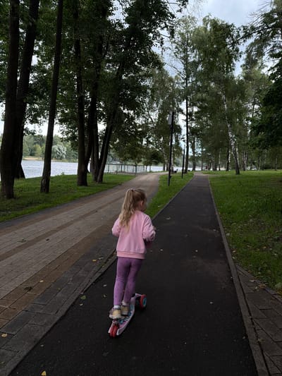 A young girl with blonde hair tied in a ponytail rides a pink scooter on a paved path in a park located in Malakhovka, Russia. She is wearing a pink sweatshirt and purple leggings. The path is next to a body of water, possibly a lake or river, and is lined with tall trees. On the opposite side of the path from the water, there is a grassy area with more trees, some of which appear to be birch trees with white bark. The sky is overcast.