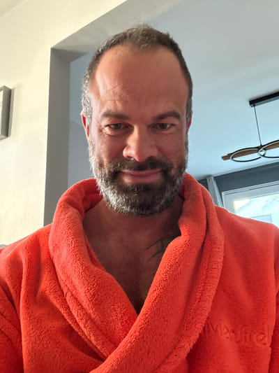 A middle-aged man is positioned at a close-up angle, wearing an orange bathrobe. He has a salt-and-pepper beard, short graying hair, and a tattoo visible on his upper chest. The man appears to be indoors, with a glimpse of a window and lighting fixtures in the background. It is not possible to determine if the image was taken in Marche-en-Famenne, Belgium, or to deduce specific actions or interactions based solely on this image. The environment consists of interior walls and ceiling, with lighting elements.