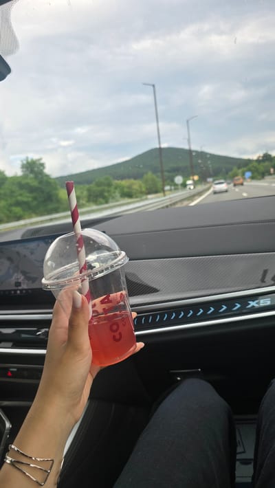 The image shows a person's hand holding a clear plastic cup with a red beverage and a striped straw, possibly inside a car. The cup has "COSTA COFFEE" printed on it. The scene outside the car window appears to be a highway with trees and hills, suggesting a travel setting. The sky is cloudy, indicating a potentially overcast day. The person is wearing a black garment and a geometric silver bracelet on their wrist. The interior of the car, with a dark dashboard featuring LED lights, is also visible. The image orientation is rotated. The location is Studena, Bulgaria.