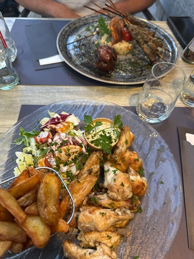 The image shows a table setting at a restaurant, likely in Durbuy, Belgium. In the foreground, a clear glass plate holds a meal of chicken wings, potato wedges, and a side salad with dressing. The chicken wings are golden brown and seasoned with herbs. The potato wedges are thick-cut and appear crispy. The salad consists of lettuce, cherry tomatoes, and red onions, topped with a creamy dressing and parsley. A small metal basket holding some of the potato wedges is also visible. In the background, another plate contains a meal of skewers with meat, possibly lamb or beef, served with rice and a piece of roasted red pepper or tomato. Two glasses of water and a bottle of what appears to be water or a soft drink are on the table. A dark placemat is under some of the dishes. The hands of two people are partially visible in the background, suggesting they are dining at the table. The overall environment appears to be outdoors, given the natural light and the wooden table surface.