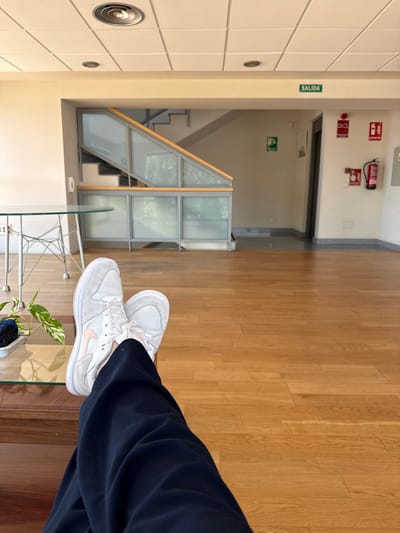 The image depicts an indoor scene with polished wooden flooring. In the foreground, a person's legs are crossed, wearing dark pants and white sneakers with pink accents. The person is sitting on what appears to be a wooden bench or furniture, with a glass-topped table in front of them. A small plant is visible on the table. In the background, a modern staircase with glass railings leads upwards. The ceiling features recessed lighting and a circular vent. On the wall in the background, there are several signs, including a green "SALIDA" (exit) sign, and red signs with pictograms, one of which indicates a fire extinguisher. The overall environment suggests an indoor waiting area or common space, possibly within a public building or institution. Given the "SALIDA" sign, the presence of fire safety equipment, and the architectural style, it is plausible this location is within Getafe, Spain, perhaps in a business, educational, or public facility.