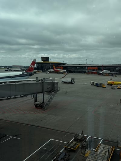 This is an eye-level, wide shot of an airport tarmac under a cloudy sky. The scene is dominated by a large passenger jet in the foreground on the left, with its boarding jet bridge attached. The jet has a white fuselage with a black stripe and a red and white tail fin bearing the logo "N". The jet bridge is gray and labeled with the number "129".
Further back, on the tarmac, several other aircraft are visible. One is a red and yellow plane with a distinct fin. Another aircraft, further to the left, is white with a blue and red tail. Various airport vehicles, including a yellow fuel truck, a white truck with "Ruzhopolnap" written on it, and several small yellow baggage tugs, are scattered across the tarmac.
In the background, a modern airport terminal building with a dark, reflective facade and a yellow roof structure is visible. A control tower with a dark, rectangular top rises above the terminal. The ground is paved tarmac with red and white markings. The overall impression is of a busy airport environment, likely during daytime, with overcast weather conditions. The location is Rzhavki 2, Russia.