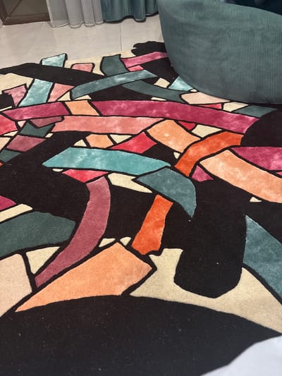 The image shows a section of a multi-colored, patterned rug with black as a prominent background color. The rug features geometric shapes in various colors including shades of pink, orange, teal, green, and beige, outlined in black. To the upper left of the rug is a folded piece of corduroy fabric in a teal color. In the lower left corner, part of a window is visible. The location is stated to be Arinj, Armenia, although this is not evident in the image itself. No people are visible.
