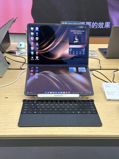 The image shows a Huawei MateBook Fold on display, with a keyboard attached. The screen displays Chinese characters and icons. A stylus labeled "HarmonyOS" is attached to the side of the device. The MateBook Fold is placed on a wooden surface. In the background, a sign with more Chinese characters is visible, alongside other devices on display. The location appears to be a store in Yuexiu District, China.