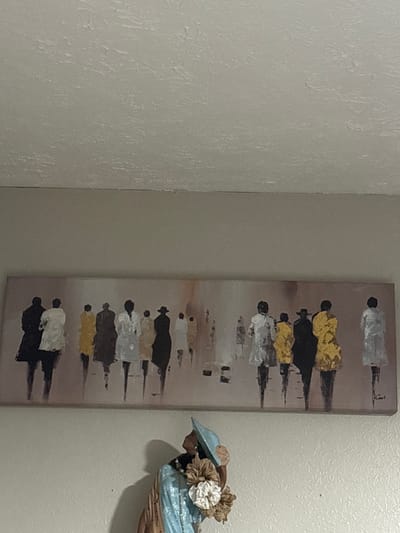 The image displays an indoor scene in Oklahoma City, United States, featuring a wall adorned with artwork and a figurine. The upper portion of the frame shows a light-colored, textured popcorn ceiling meeting a light beige, smooth wall. Centrally positioned on the wall is a large, rectangular, horizontally oriented abstract painting. This painting depicts numerous stylized human figures, appearing elongated and in motion, rendered with minimal detail against a muted beige and light brown background. The figures are clothed in varying colors, including black, white, light gray, yellow, and light brown; some wear hats. The artist's signature, "Cool," is visible in the bottom right corner of the canvas. Partially visible in the lower-center foreground is a decorative figurine depicting an African American woman from the bust up. She wears a light blue broad-brimmed hat, a light blue top with a textured surface, and a necklace, accessorized with light-colored, possibly fabric, floral embellishments near her right shoulder.