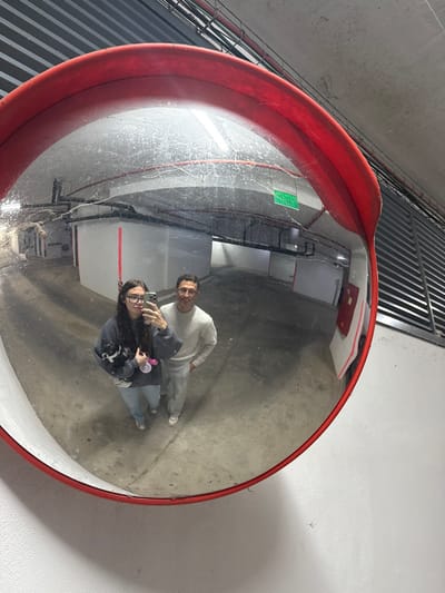 A wide-angle, slightly rotated image captures two individuals taking a selfie reflected in a large, circular convex mirror. The mirror, framed in red, is mounted on a light-colored wall.
In the reflection, a woman is positioned slightly in front, holding a smartphone in her right hand to take the picture. She wears glasses, a dark hooded top, light-colored jeans, and white sneakers. Her long, dark hair falls over her shoulders. She has a playful expression, with her tongue slightly extended. Her left arm cradles a small black and white dog, which is also looking towards the camera. Beside her, a man with short dark hair and a beard is smiling, looking towards the camera. He is dressed in a light-colored (possibly white or light grey) long-sleeved top, matching pants, and white sneakers.
The mirror's surface shows signs of wear, with numerous small scratches, dust, or dirt specks visible across its curvature. The reflected environment suggests an indoor setting, likely a parking garage or industrial area, with a concrete floor, light-colored walls, and some visible piping or conduits. In the upper left quadrant of the reflection, a small green rectangular sign with white text is visible on a wall; the text is too indistinct to read. A red panel or box is also reflected higher up on a wall in the background.
Outside the mirror's frame, to the upper left of the image (due to the rotation), a large metal grate or ventilation system with parallel bars is visible against a darker background. To the upper right, the plain light-colored wall on which the mirror is mounted is visible, with subtle shadows. The image itself is rotated counter-clockwise, giving the impression that the wall is positioned diagonally.