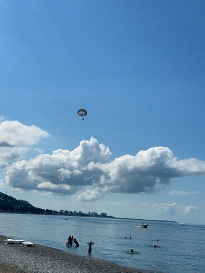 A wide-angle, vertical, eye-level shot captures a clear blue sky over a calm sea and a pebbled beach. In the sky, a parasail with a colorful canopy is being towed by an unseen boat, carrying two individuals. Puffy white clouds are scattered across the sky, with a larger formation of clouds nearer to the horizon.
The sea is a tranquil blue, with a gentle shoreline. Several people are visible in the shallow water near the beach, some wading and others swimming. One person stands in the water with their arms raised, while another group appears to be gathered together. In the distance, a motorboat is chugging across the water, leaving a small wake.
Along the distant coastline, a city skyline with numerous buildings is visible, suggesting an urban area. A green, tree-covered hill slopes down towards the city on the left side of the image. The beach in the foreground is made up of small, gray pebbles, with a few empty white lounge chairs placed on it. The overall scene depicts a leisurely summer day at the beach, with recreational activities taking place on both the water and in the air. The location is Abaata, Abkhazia.