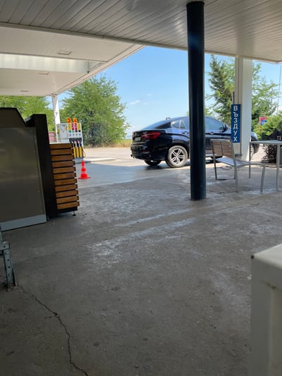 The image depicts a gas station in Gorni Dabnik, Bulgaria, under a covered canopy. A dark-colored SUV is parked near the pumps. To the right of the vehicle, a blue sign with Cyrillic text "ВЪЗДУХ" (meaning "AIR") is visible. A metal bench with wooden slats is positioned near a support column. In the foreground, a concrete surface shows signs of wear and a crack. In the background, trees and a clear blue sky are visible. A bright orange traffic cone is placed on the ground near the gas pumps. The overall scene suggests a typical operation of a gas station.