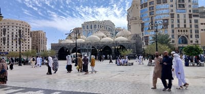 This image depicts a bustling outdoor plaza in Medina, Saudi Arabia, specifically in front of the Al-Ghamamah Mosque, which is located southwest of the Prophet's Mosque (Masjid an-Nabawi) in the city center.

The scene features a wide, light-colored paved square under a partly cloudy blue sky. In the mid-ground, the Al-Ghamamah Mosque stands prominently, characterized by its dark stone facade, numerous arched entrances, and a roof adorned with multiple white, low-profile domes. Surrounding the plaza are several multi-story modern buildings, likely hotels or commercial establishments, with varying architectural styles, including facades with reflective blue windows and others with more traditional light-colored stonework. A distinct minaret-like structure is visible on the far right.

Approximately 50 to 70 individuals are visible across the plaza. The majority are adults dressed in traditional Islamic attire: men wear thobes in white, brown, or dark colors, with some sporting white head coverings, while women wear abayas and headscarves, some in solid dark colors, others with patterns. Most people are walking, moving in various directions across the paved area. Some groups are standing, appearing to converse, while a few individuals are seated on stone benches or low wall features. In the immediate foreground on the right, three adult males are walking towards the left; one wears a light brown thobe, another a dark blue thobe, and the third a white thobe with a white head covering. A white vehicle, possibly an SUV, is partially visible in the mid-ground behind a cluster of people near the mosque. Ornate black street lamps are positioned throughout the plaza. Dark bird-like figures are seen flying against the sky in the mid-ground. Shadows cast by individuals and structures suggest daylight conditions.