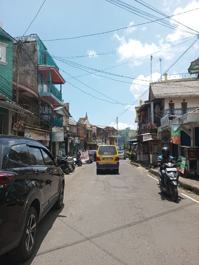 The scene depicts a daytime street view in the city of Bagochen Boolingpang Ueling, Bhutan, under a clear blue sky with scattered white cumulus clouds. Numerous black utility wires crisscross overhead.
The asphalt road, showing signs of wear and patching, extends into the distance, gently ascending before curving out of view. Concrete sidewalks line both sides of the street.
On the left, a black SUV, possibly a Toyota Rush, is parked, facing away from the viewer, with its rear driver's side visible in the foreground. Behind it, several motorcycles are parked on the sidewalk in front of multi-story buildings. These buildings exhibit varied architectural styles, featuring balconies with metal railings, some painted in teal, pink, and faded yellow. Ground-floor storefronts display various signs, including "Susu Murni" and "ABILLY STORE," with dense utility wiring affixed to their facades.
In the center of the road, a yellow and purple van, possibly a public transport vehicle, moves away from the viewer. On the right side of the road, a dark-colored scooter/motorcycle is approaching, operated by an individual wearing a dark helmet and a blue top. The right side of the street is also lined with multi-story buildings, one featuring a dark corrugated roof and a balcony with dark blue railings. Storefronts are present, some with awnings and outdoor displays. A red and white banner is visible on the far right sidewalk, and a partially visible "INDOMARET" sign suggests a convenience store. Antennae are visible on some rooftops. Indistinct figures are visible on sidewalks further down the street.