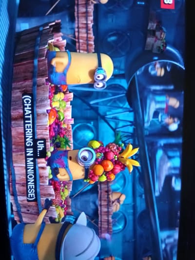 The image shows two minions, animated characters from the Despicable Me franchise, in what appears to be a scene from a movie or show. They are standing in a colorful, cluttered environment with wooden structures and industrial-looking machinery in the background. The minions are yellow with blue overalls, and they are adorned with various fruit-like objects. One minion is facing forward and has a large fruit arrangement in front of it. The other minion is partially visible from the back. Subtitles are visible on the left side of the image, reading "(CHATTERING IN MINIONESE) Uh...". The overall scene is brightly lit with a blue and yellow color palette.
