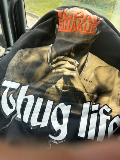 The image shows a black sweatshirt with a print of Tupac Shakur. The word "THUG" is printed vertically in white letters. The print includes Tupac's image, his name, and the dates 1971-1996. The sweatshirt is lying on the dashboard of a vehicle, possibly in the city of Celle, Germany, based on visual markers of a car interior and background elements.