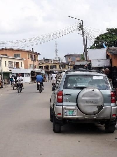 The scene depicts an urban street in Alaiyabiagba, Nigeria, under an overcast sky. A light gray Toyota RAV4 SUV, bearing Nigeria license plate KSF 211GF, is parked on the right side of a paved asphalt road, viewed from the rear. The vehicle has a roof rack and a spare tire mounted externally on the tailgate, its cover showing signs of wear.
In the midground, two individuals are operating motorcycles on the road. One person, wearing a blue shirt, rides a motorcycle away from the viewer. Another person, in a white shirt, rides a motorcycle pulling a small wheeled cart towards the viewer on the left side of the road. Several other pedestrians are visible walking further down the street on the left.
The left side of the street features a white, kiosk-like structure with a green roof. Behind it and extending into the background are numerous buildings, including single-story commercial stalls and multi-story residential or mixed-use structures painted in various muted colors such as peach, cream, and brown. On the right, a wooden utility pole with a street light fixture is heavily laden with multiple tangled electrical wires. Further right, a person with a shaved head is partially visible standing near some structures.
In the background, a tall, lattice-style telecommunications tower stands prominently. Green foliage from trees is visible on the far right.