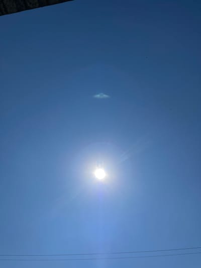 The image depicts a clear blue sky with the sun prominently visible, emitting bright rays and a lens flare. Above the sun, a small, hazy cloud formation is present. In the upper left corner, a dark, textured object, possibly the edge of a building or structure, juts into the frame. In the distance, faint horizontal lines, likely power or telephone wires, are visible near the bottom of the image. A few tiny dark specks, possibly birds or dust, are scattered throughout the sky. The scene is set in Kryzhanivka, Ukraine, under daylight conditions.