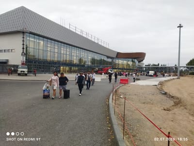 This is a photograph taken at an airport in Bishkek, Kyrgyzstan. Two women are walking on a paved area, each pulling a suitcase. They are wearing casual clothing: one in a pink long-sleeved shirt and white shorts, the other in a dark blue jacket and a grey skirt. In the background, a modern airport terminal building with large glass windows is visible. The building has a distinctive grey, diamond-patterned roof. Several other people are walking around the airport, and vehicles, including a white truck and a red crane, are present. The scene appears to be a busy area outside the airport, possibly a drop-off or pick-up zone. The sky is overcast, suggesting cloudy weather. The timestamp in the lower right corner indicates the photo was taken on July 2, 2026, at 18:55, indicating it was taken in the early evening. The text "SHOT ON REDMI 9T" is visible in the lower left corner. There is also construction work in progress in the foreground, with red tape and construction materials visible.