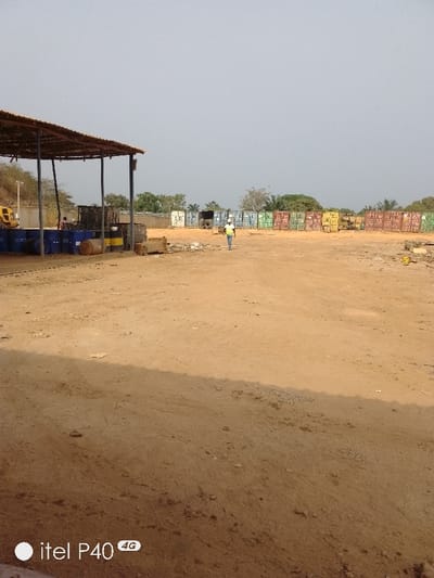 An outdoor industrial or storage yard in Hoche, Nigeria, is depicted under a hazy, light gray sky. The foreground and midground consist of a vast expanse of reddish-brown dirt, showing signs of vehicle traffic and scattered debris.

On the left side of the midground, a large, open-sided shelter with a corrugated metal roof is present, supported by dark poles. Beneath this shelter, several blue and yellow industrial barrels or drums are visible, some stacked and some lying horizontally. A partial view of yellow heavy machinery, possibly an excavator, is visible to the left behind the shelter. A person wearing a red shirt is partially visible under the shelter, near the barrels.

In the center of the midground, a person walks from left to right across the dirt. This individual is wearing a white hard hat, a yellow-green high-visibility vest over a light-colored shirt, and dark pants.

The background features an extensive line of various colored shipping containers (white, light blue, green, yellow, and rust/brown) arranged in rows across the entire width of the frame. Behind these containers and in the far distance, sparse vegetation and trees are visible. A fence is partially visible behind the shelter and some containers. A utility pole stands on the far left. The overall scene suggests a commercial or industrial storage operation.